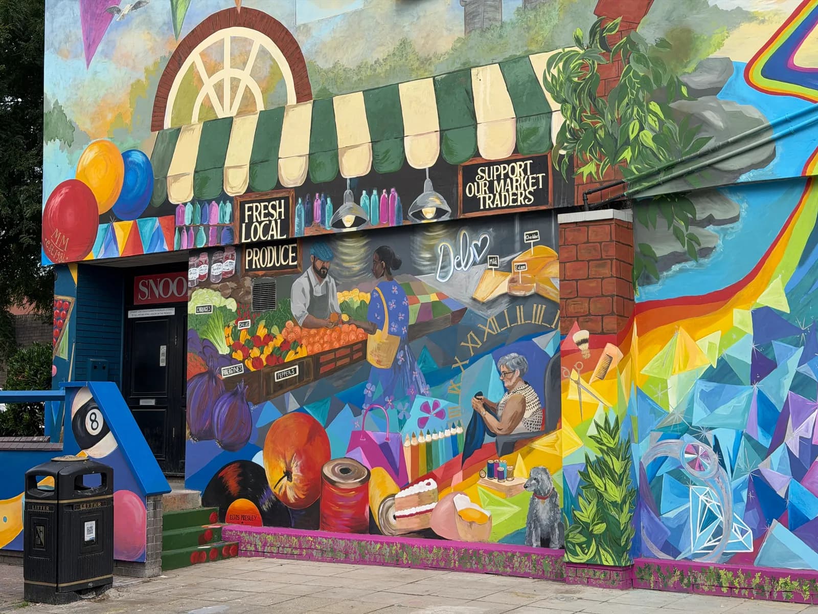 A vibrant and detailed mural on the side of a building depicts a market scene with a fruit stall, a woman sewing, and various colorful geometric patterns and objects.