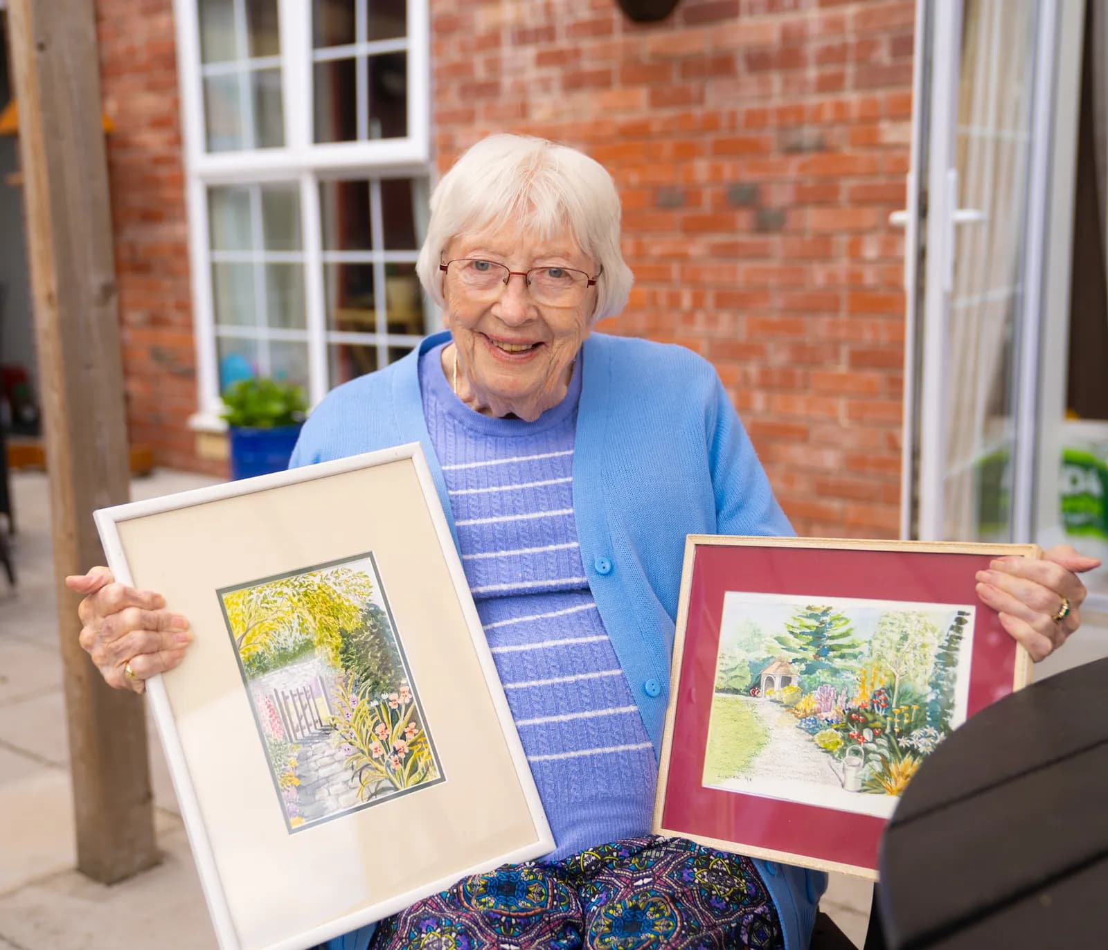 An elderly woman with white hair and glasses smiles while holding two framed watercolor paintings of gardens.