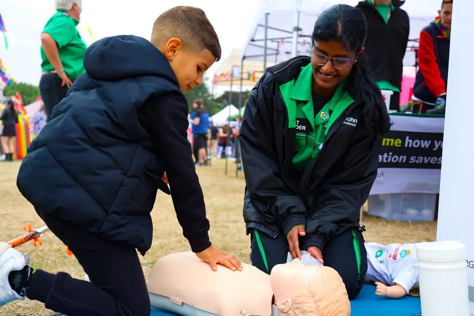 A young boy practices CPR on a mannequin while a St. John Ambulance volunteer in a green and black uniform watches and assists.