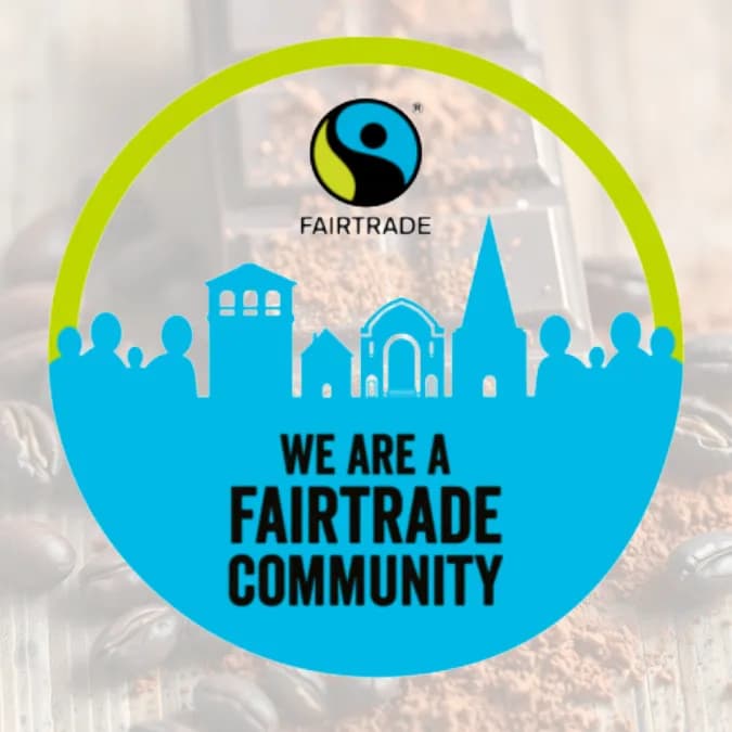 A circular logo with a green border and light blue interior, featuring silhouettes of buildings and people under the Fairtrade logo, with the text 'WE ARE A FAIRTRADE COMMUNITY' at the center, all overlaying coffee beans and ground coffee.